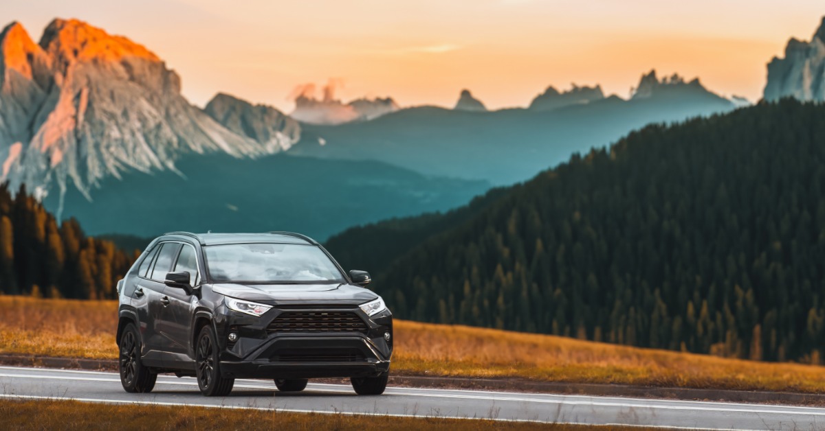 A black SUV drives down a road as the sun sets. Mountains and a forest of trees are in the background.