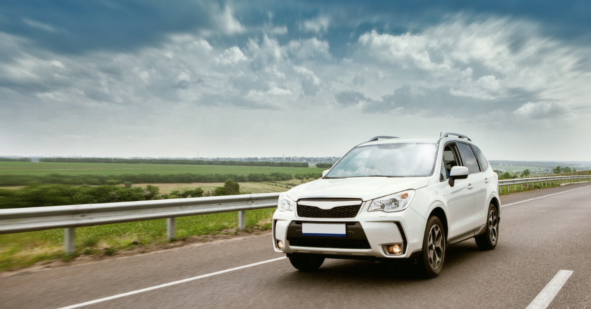 A white SUV drives down a road with farm fields blurred in the distance. The sky is dark and cloudy.