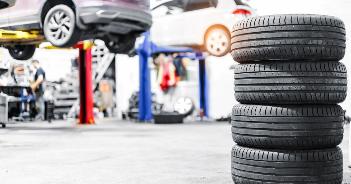 A stack of four worn tires sits on the concrete floor of an auto shop. Cars are on lifts in the background.
