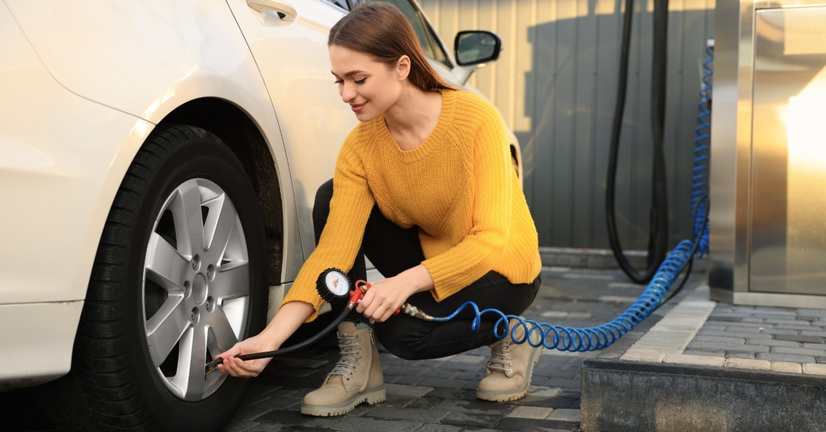 A woman holding a blue air hose kneels next to the back tire of a white van. She holds the hose against the tire.