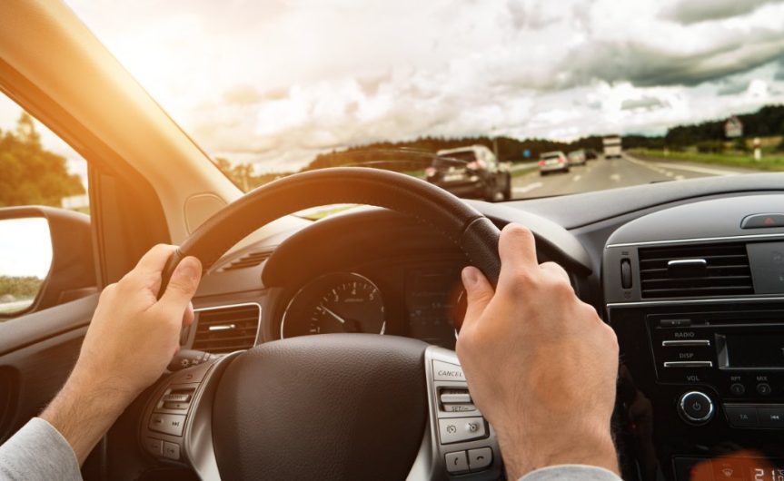 A point-of-view of a driver holding the steering wheel of a modern car with both hands and driving on a highway.
