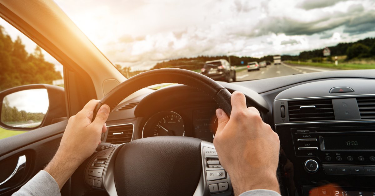 A point-of-view of a driver holding the steering wheel of a modern car with both hands and driving on a highway.