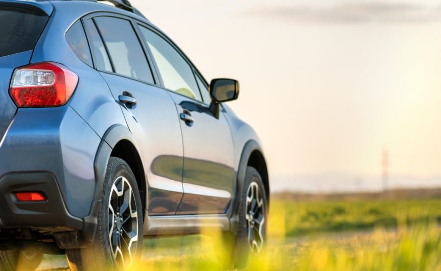 A blue car is driving down a gravel road in a rural area. There are no other cars or people on the road.