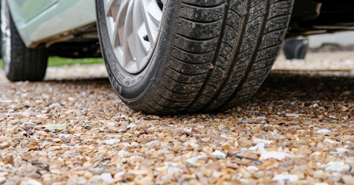 A car parked on a gravel road with small rocks of various colors underneath the tire. The tire has dirt and minor marks.