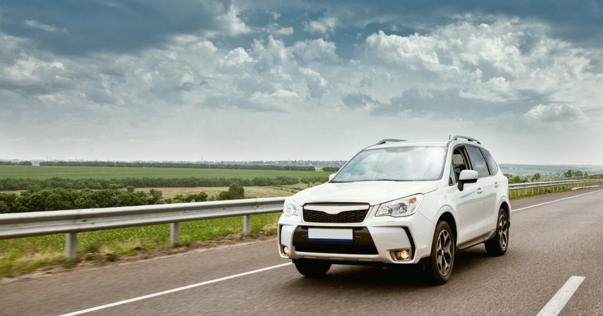 A modern white car driving down an empty road with open fields behind it. There are dark clouds in the sky.