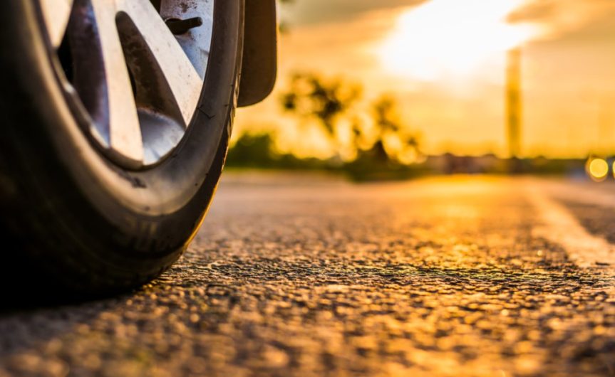 A close-up of a tire on an asphalt road. The sun peeks through a break in the clouds ahead of the tire.