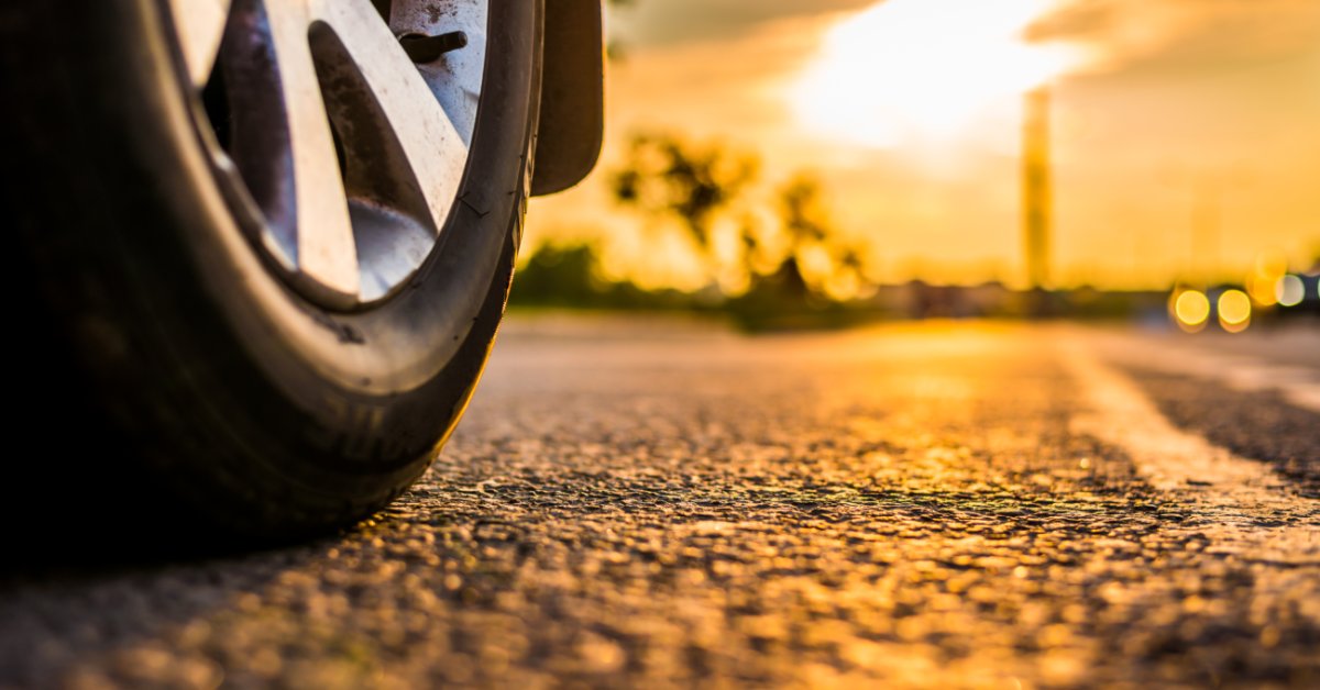 A close-up of a tire on an asphalt road. The sun peeks through a break in the clouds ahead of the tire.