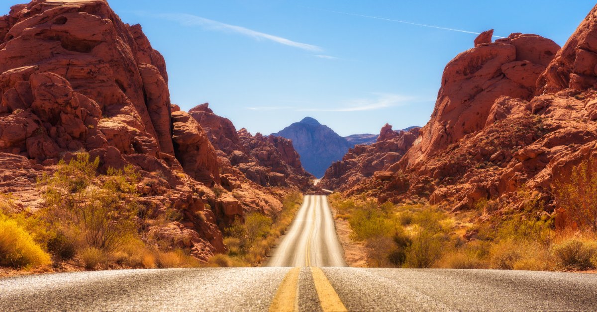 A straight desert road leading through a red rock canyon, with brush, distant mountains, and a bright blue sky.