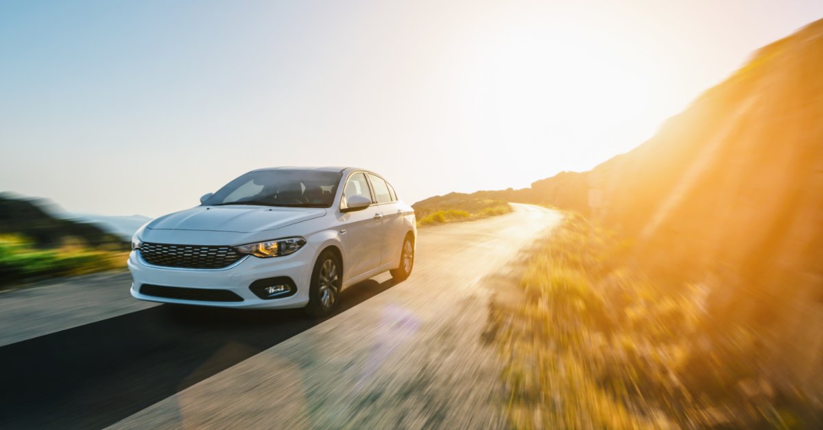 A white car drives down a windy road with the sun shining behind it. The road around the car is blurry.