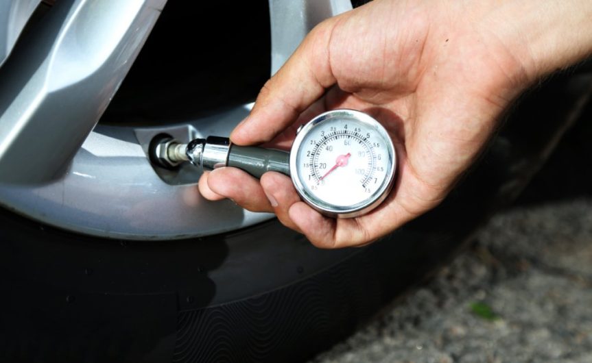 A person's hand holding a pressure gauge to a car tire to measure the pressure. The measurement shows 0 PSI.