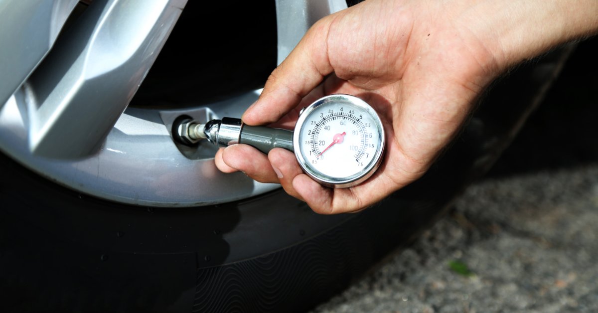 A person's hand holding a pressure gauge to a car tire to measure the pressure. The measurement shows 0 PSI.