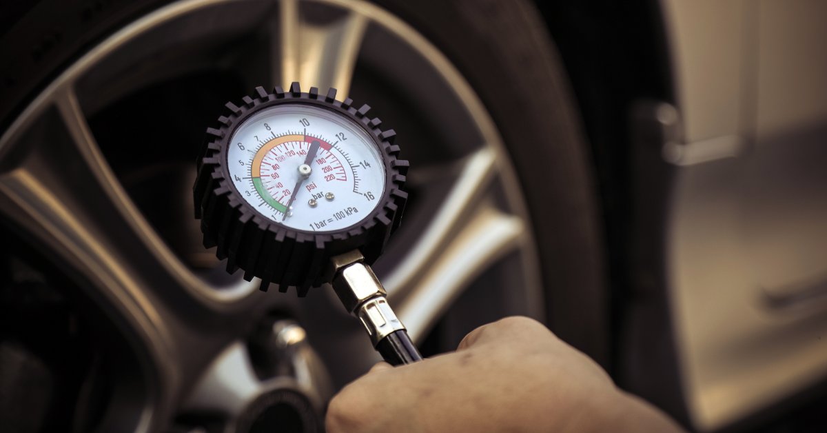 A hand holding a tire pressure gauge in front of a car tire. The gauge shows the PSI range the tire should be at.