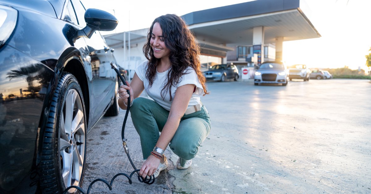 A woman at a gas station filling the tire of her vehicle with air from a hose. She is kneeling next to the vehicle.