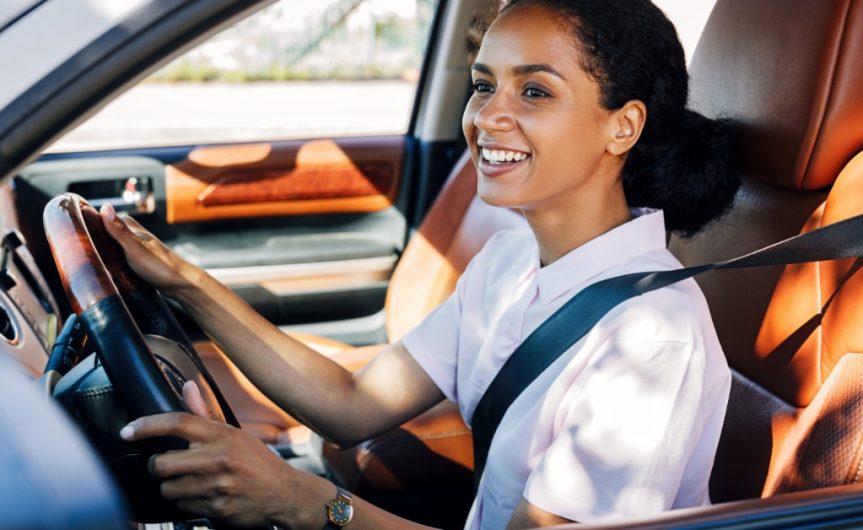 A smiling young woman driving a car with the driver's window down. She has both hands on the wheel and her seatbelt on.
