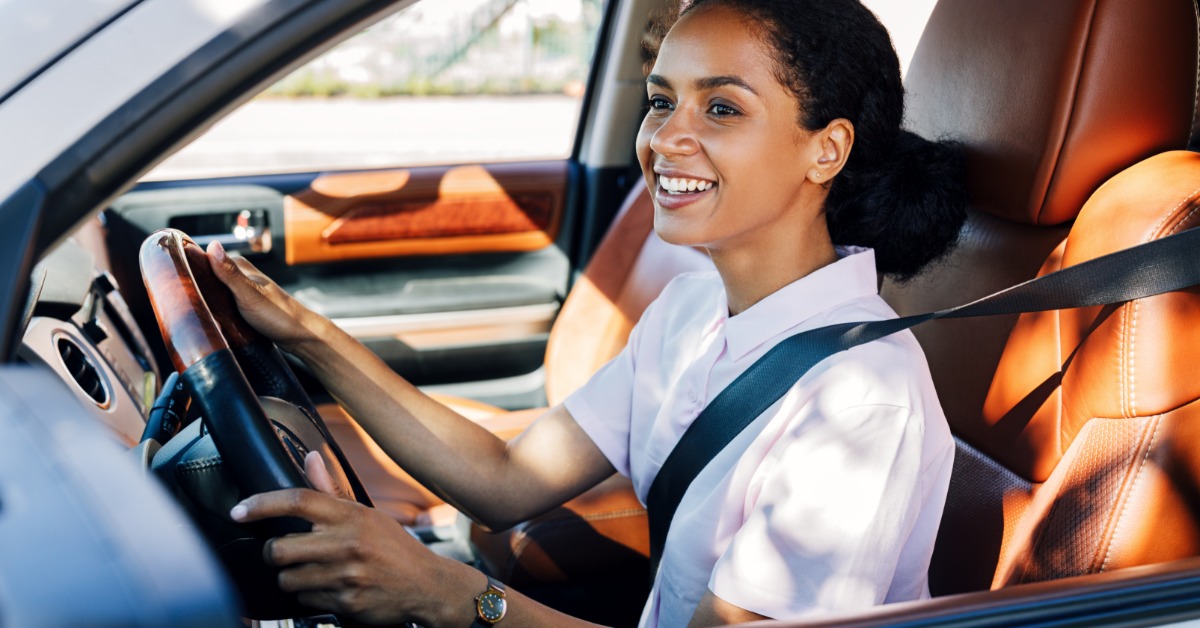 A smiling young woman driving a car with the driver's window down. She has both hands on the wheel and her seatbelt on.
