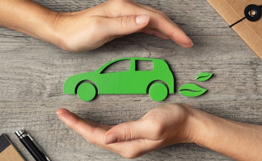 A green cutout icon of a car with leaves behind it on a wooden table, with two cupped hands above and below the car.