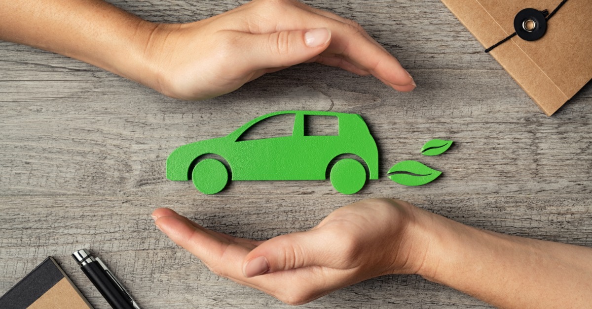 A green cutout icon of a car with leaves behind it on a wooden table, with two cupped hands above and below the car.
