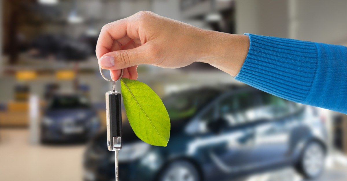 A close-up of a hand holding out a key ring with one key fob for a car on it and a green leaf. Blurred behind are cars.