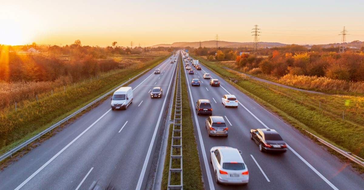 An aerial view of a highway with two lanes on either side. Several cars drive along the road with the sun setting.