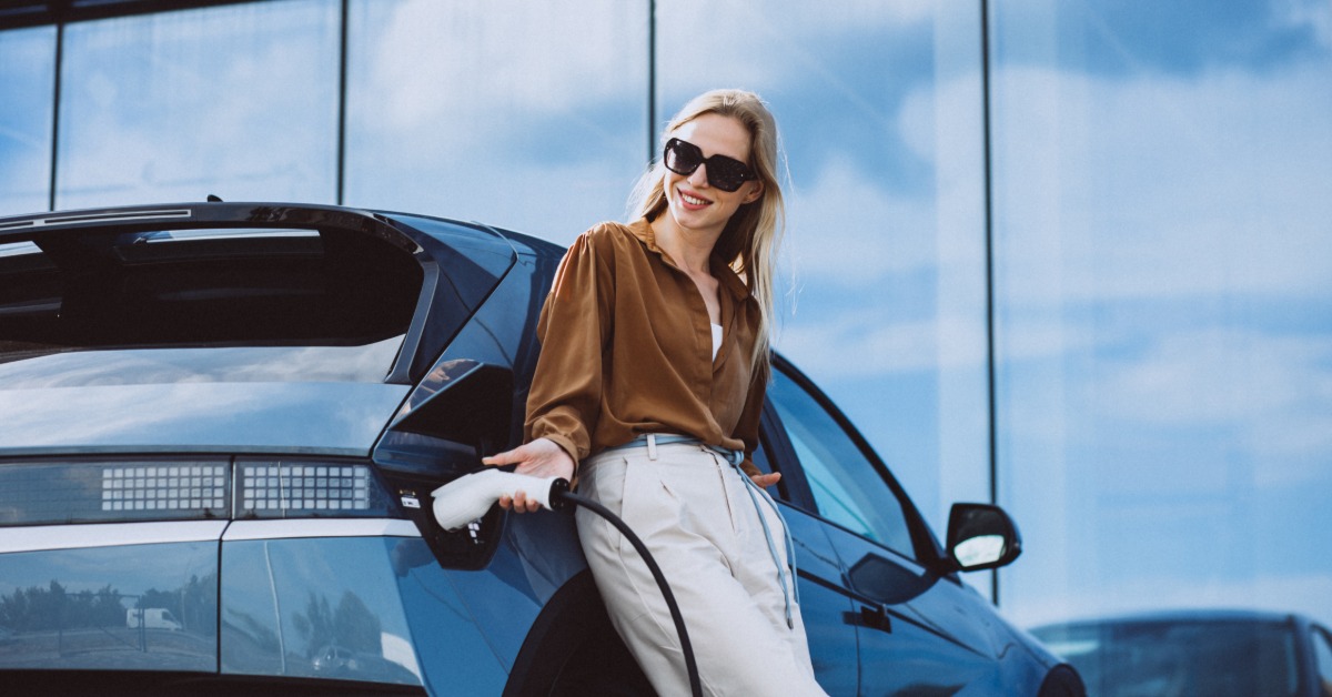 A well-dressed woman leaning against her electric vehicle while reaching to unplug the charger from the car.