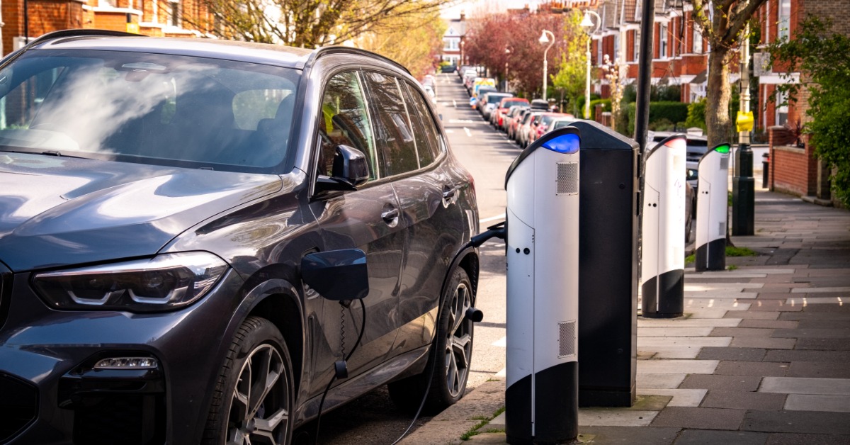 An electric car parked on a residential street next to a charging station. The cord is plugged into the vehicle.