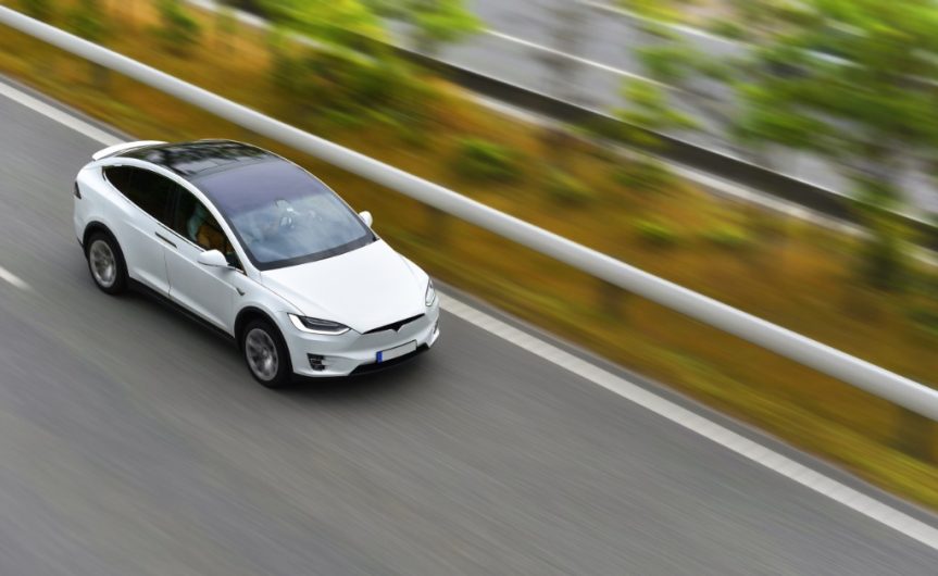 A white electric car is driving down an empty highway. The background is blurred due to the car's speed.