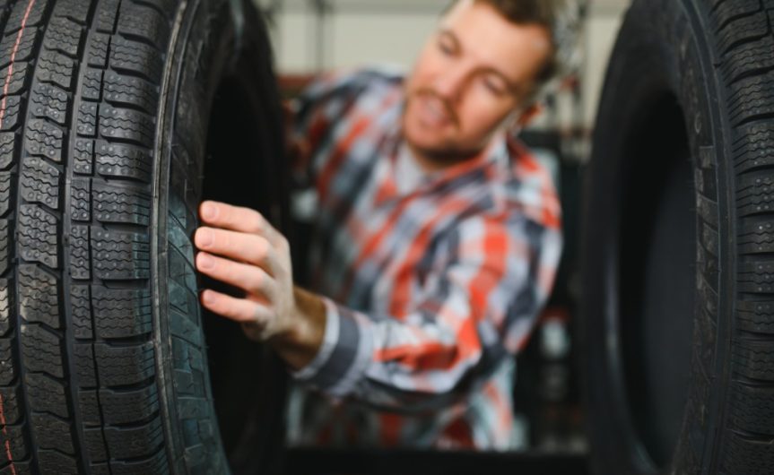 A young man in a flannel shirt reaches between a rack of new car tires to touch and read the sidewall of a tire.