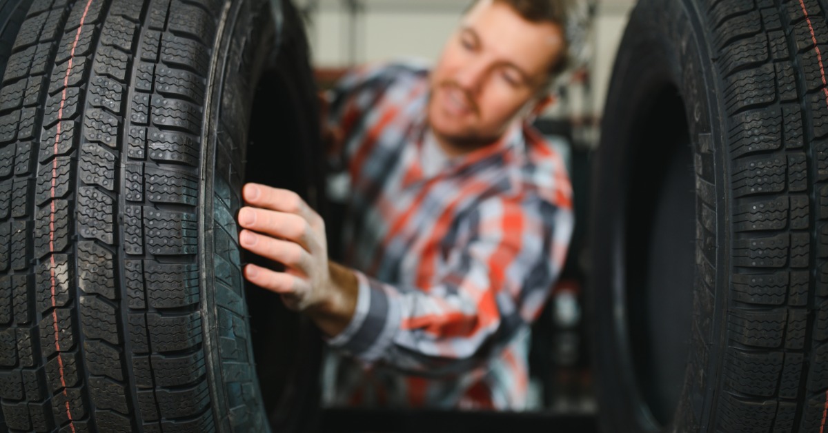 A young man in a flannel shirt reaches between a rack of new car tires to touch and read the sidewall of a tire.
