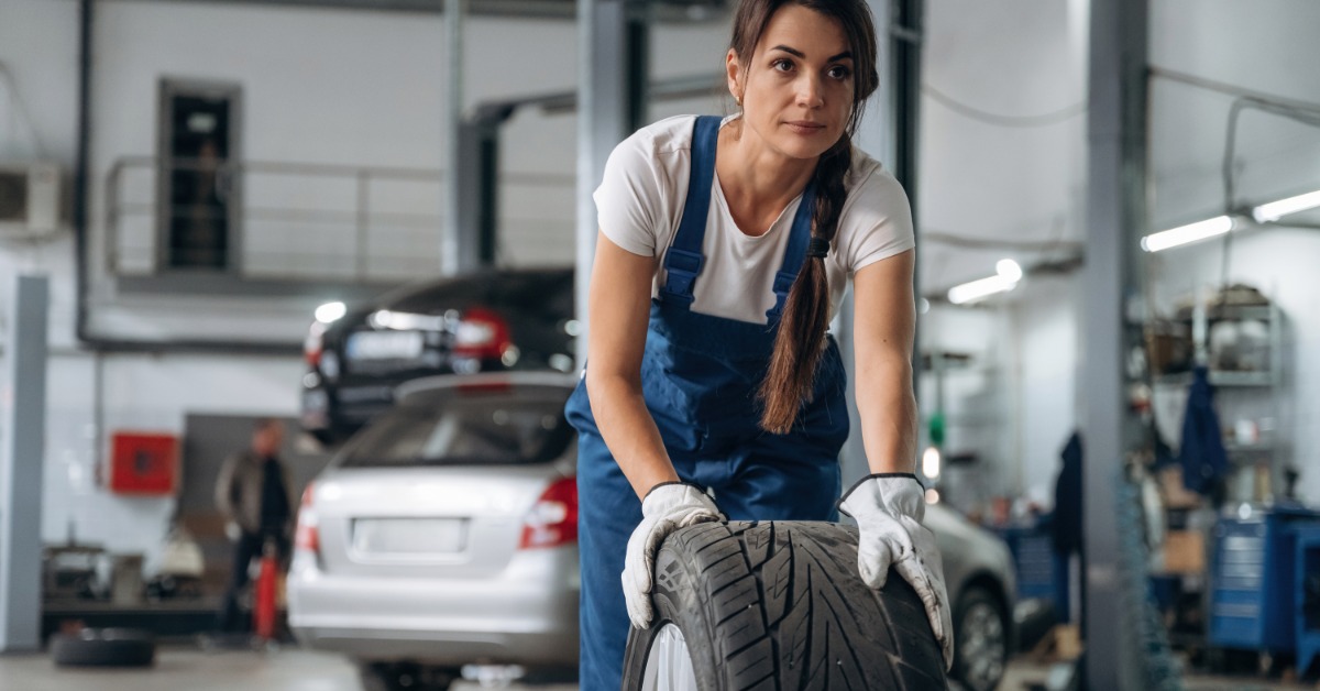 A woman in blue overalls with white gloves rolls a used tire on the floor with cars in the repair shop behind her.