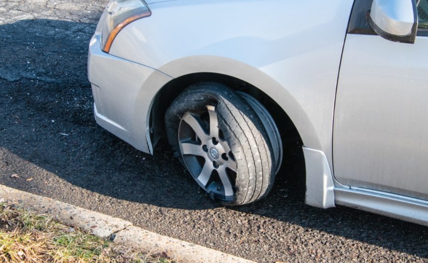 The front of a silver car with the tire on the driver's side completely blown out. The wheel and tire rim are visible.