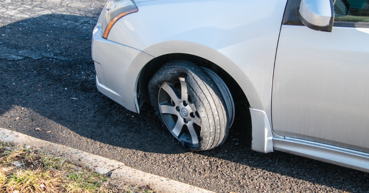 The front of a silver car with the tire on the driver's side completely blown out. The wheel and tire rim are visible.