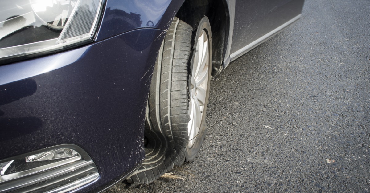 A close-up of a dark blue car on a road. The car's front tire is completely blown out, with the rubber sliding off.