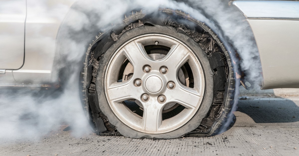 A close-up of a blown-out tire on a vehicle with the internal structure visible. Smoke surrounds the tire.