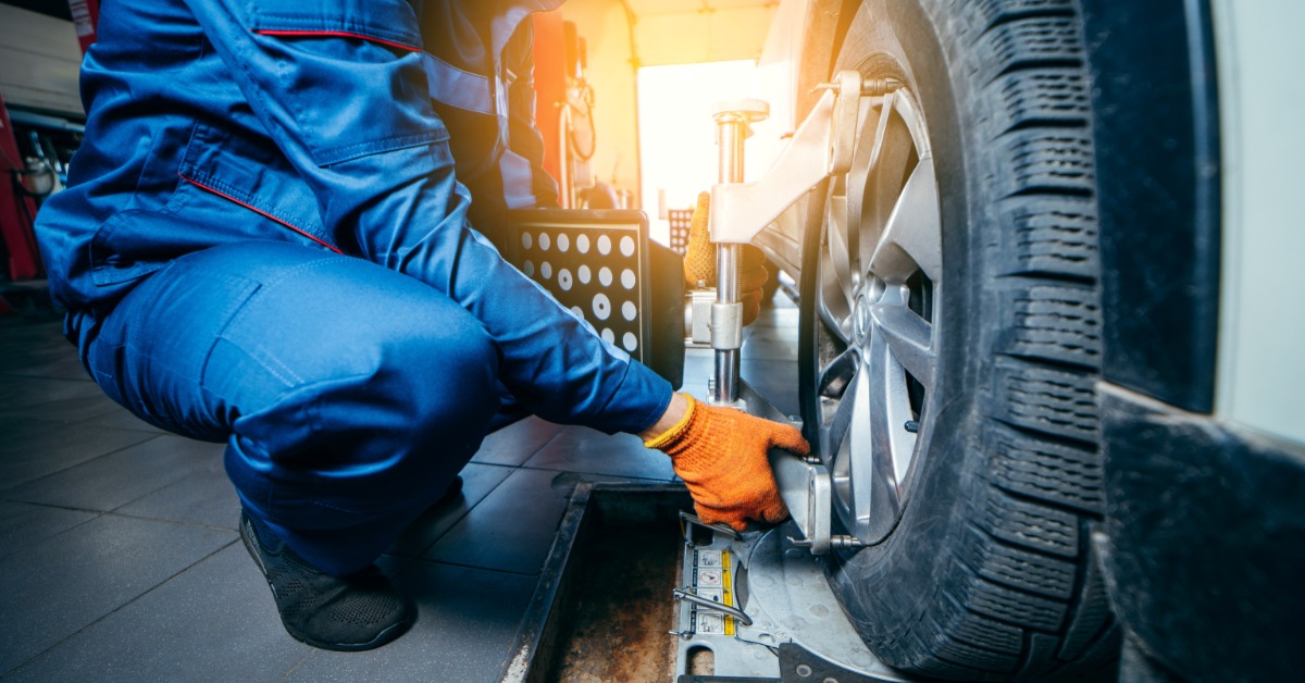 A close-up of a man in a blue jumpsuit with orange gloves crouching to install a sensor on a car for a wheel alignment.