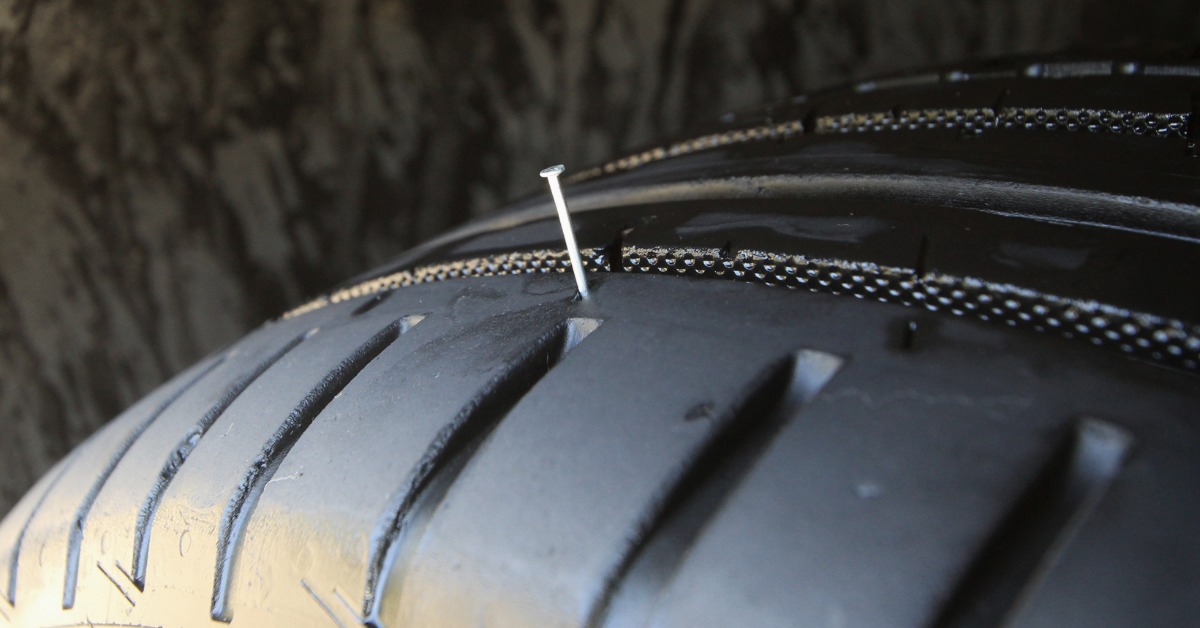 A close-up of a tire and its tread. A small nail punctures and sticks out of the tire near the tread.