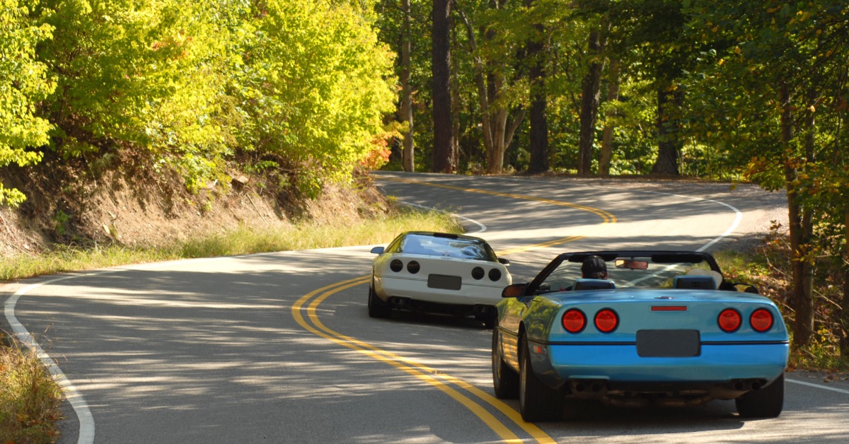 A rear view of two classic white sports cars driving on a winding road surrounded by forests on a sunny day.