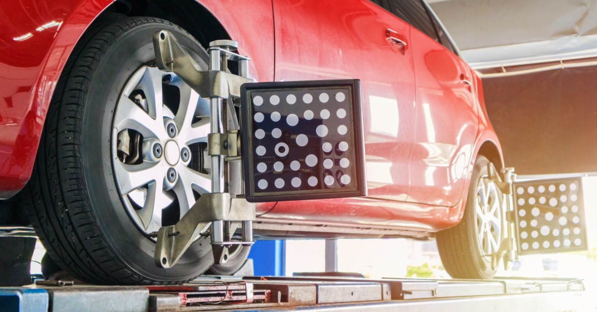 A red car in an auto shop. The front and rear wheels on one side have wheel alignment sensors clamped to them.