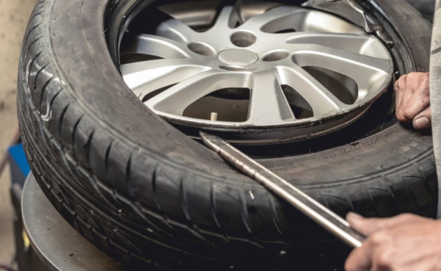 A close-up of a man using a tire bead breaker and a steel rod to remove the tire from a wheel rim disconnected from a car.