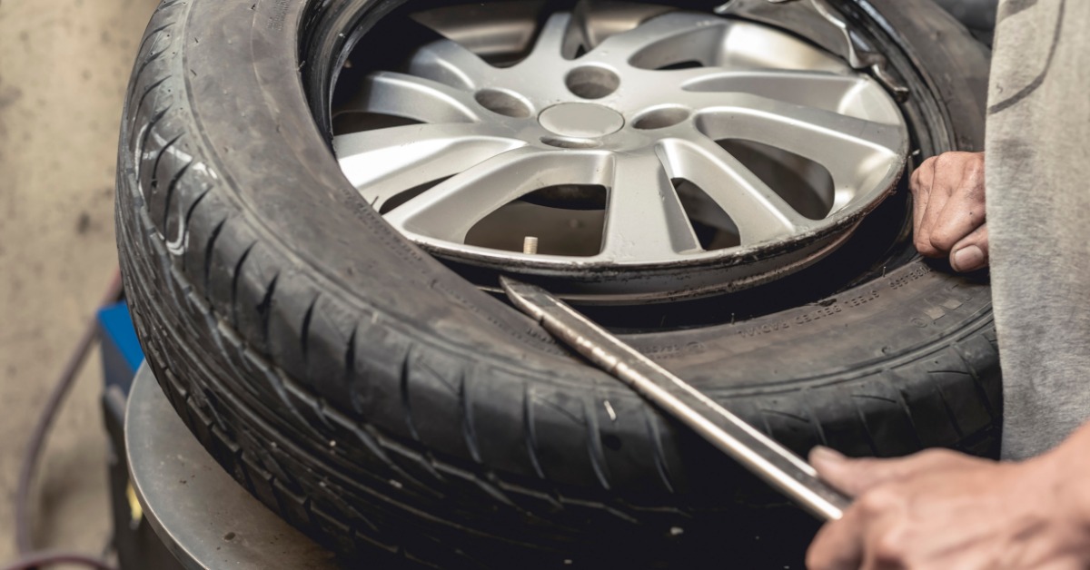 A close-up of a man using a tire bead breaker and a steel rod to remove the tire from a wheel rim disconnected from a car.