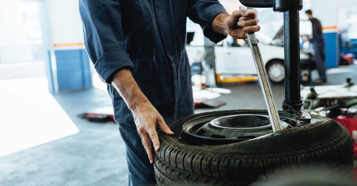 A man in a blue jumpsuit in an auto repair shop uses a steel rod to remove the tire from the rim of a wheel.