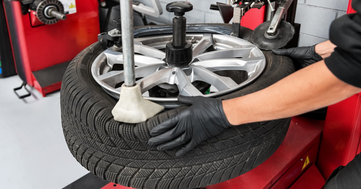 A close-up of a man with black gloves using a red tire bead breaker to remove the tire from a wheel rim.