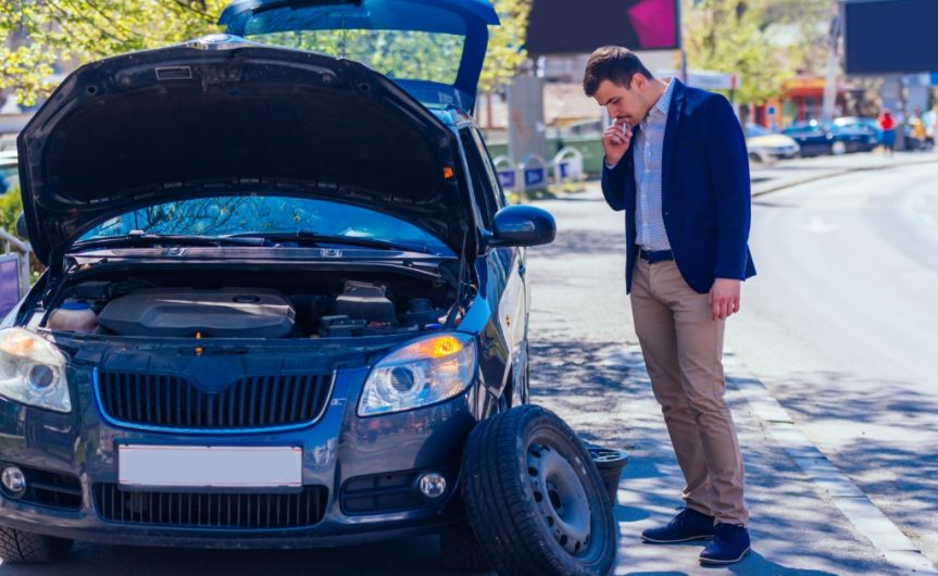 A well-dressed man looking down at his tire which is detached from the vehicle. The hood of his car is also up.