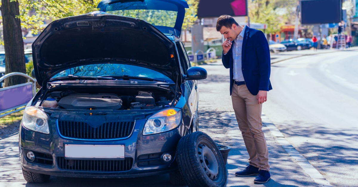 A well-dressed man looking down at his tire which is detached from the vehicle. The hood of his car is also up.