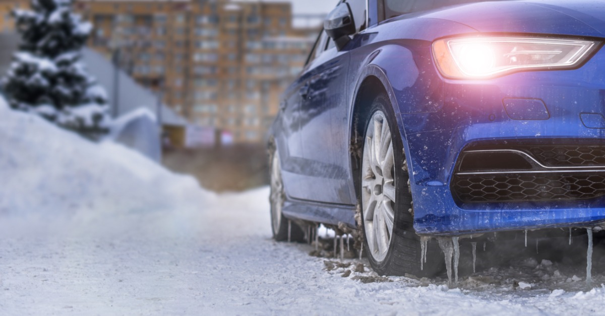 A car in the winter with the headlights on and snow on the ground around it. The bottom of the car has icicles.