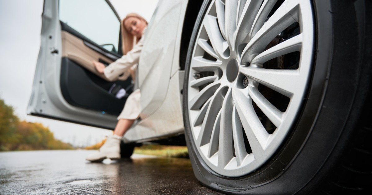 The flat tire of a white car on the side of the road. The driver is looking at the tire from the open driver's door.