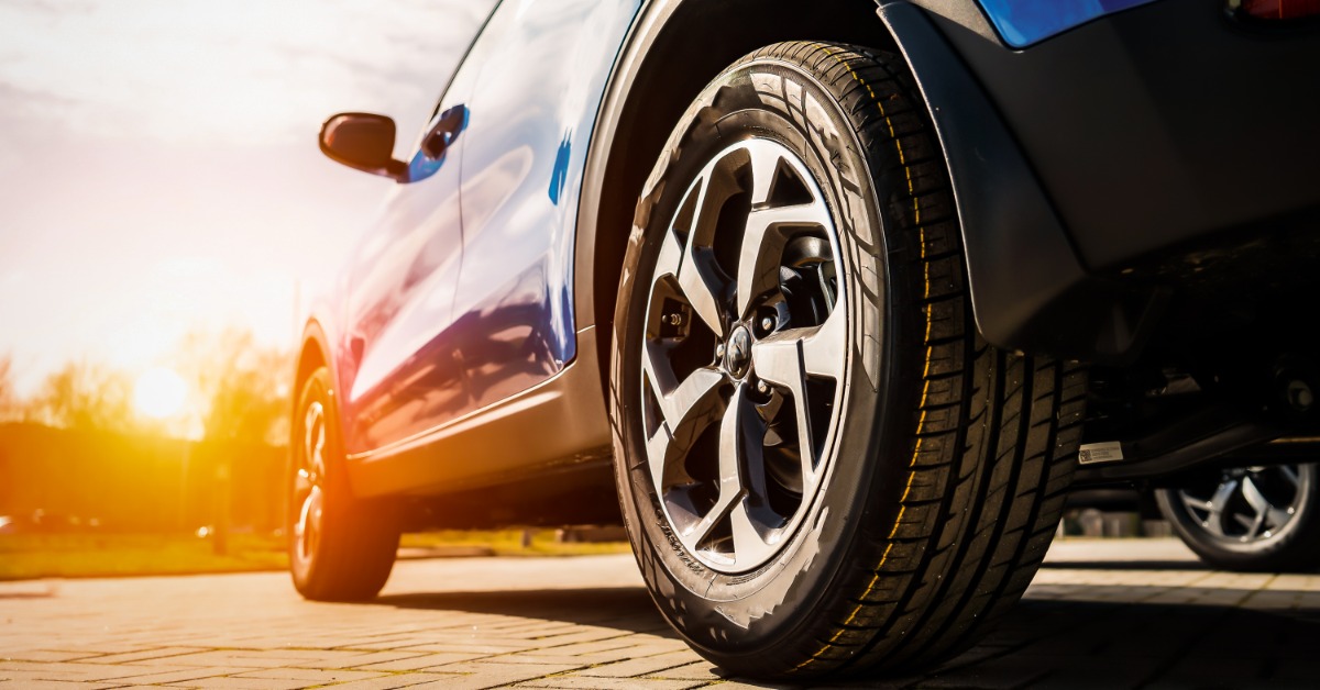 A close-up of the back and front left tire of a blue SUV. The sun peeks over a few trees in the distance.