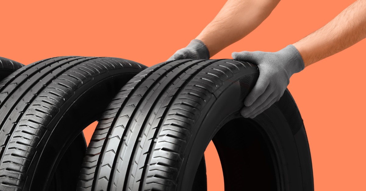 A pair of gloved hands holding three black car tires with visible tread against a solid orange background.