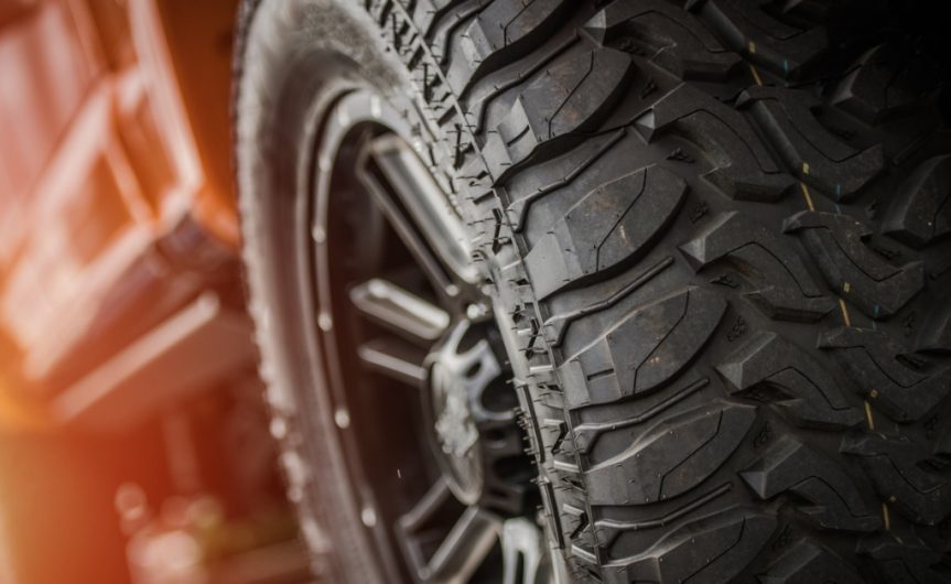 A close-up of the heavy-duty off-road tires and wheels of an orange pickup truck with a lift kit at sunset.