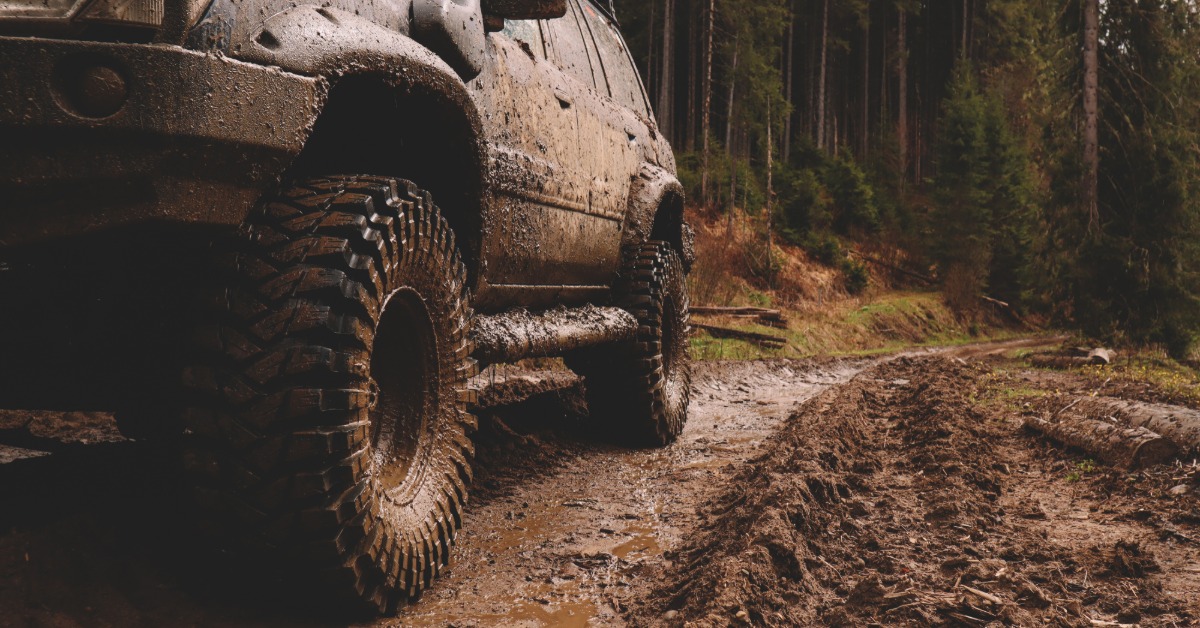 A view from the ground of an SUV with a lift kit and off-road tires parked on a muddy forest trail and covered in mud.