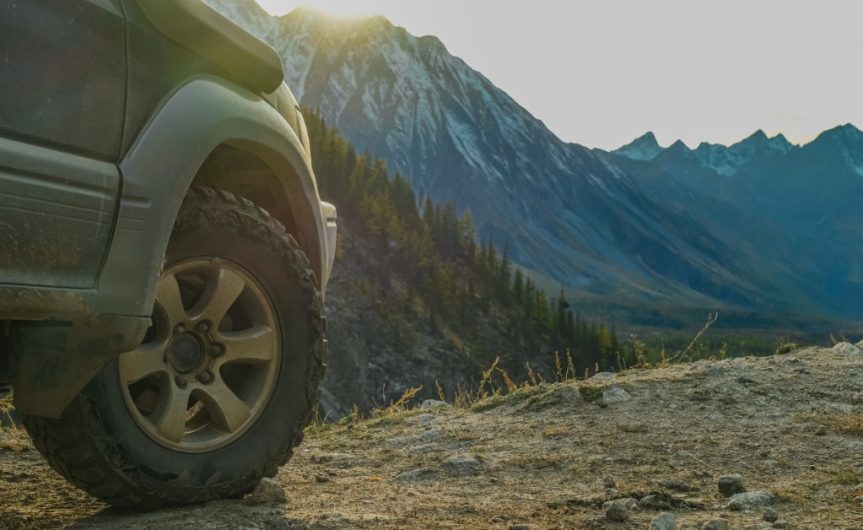 A close-up of a vehicle's front off-road tire on top of rugged terrain. A large mountain looms in the background.